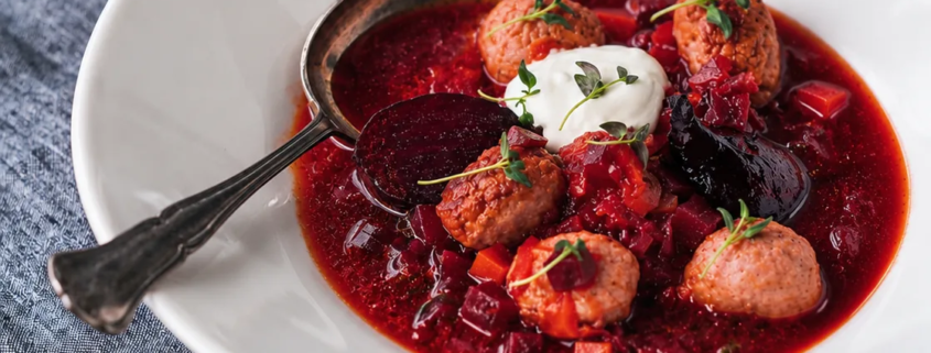 Siskonmakkara borssikeitto valkoisessa kulhossa, punajuuria, meheviä siskonmakkarapalleroita, smetanaa ja tuoreita yrttejä.